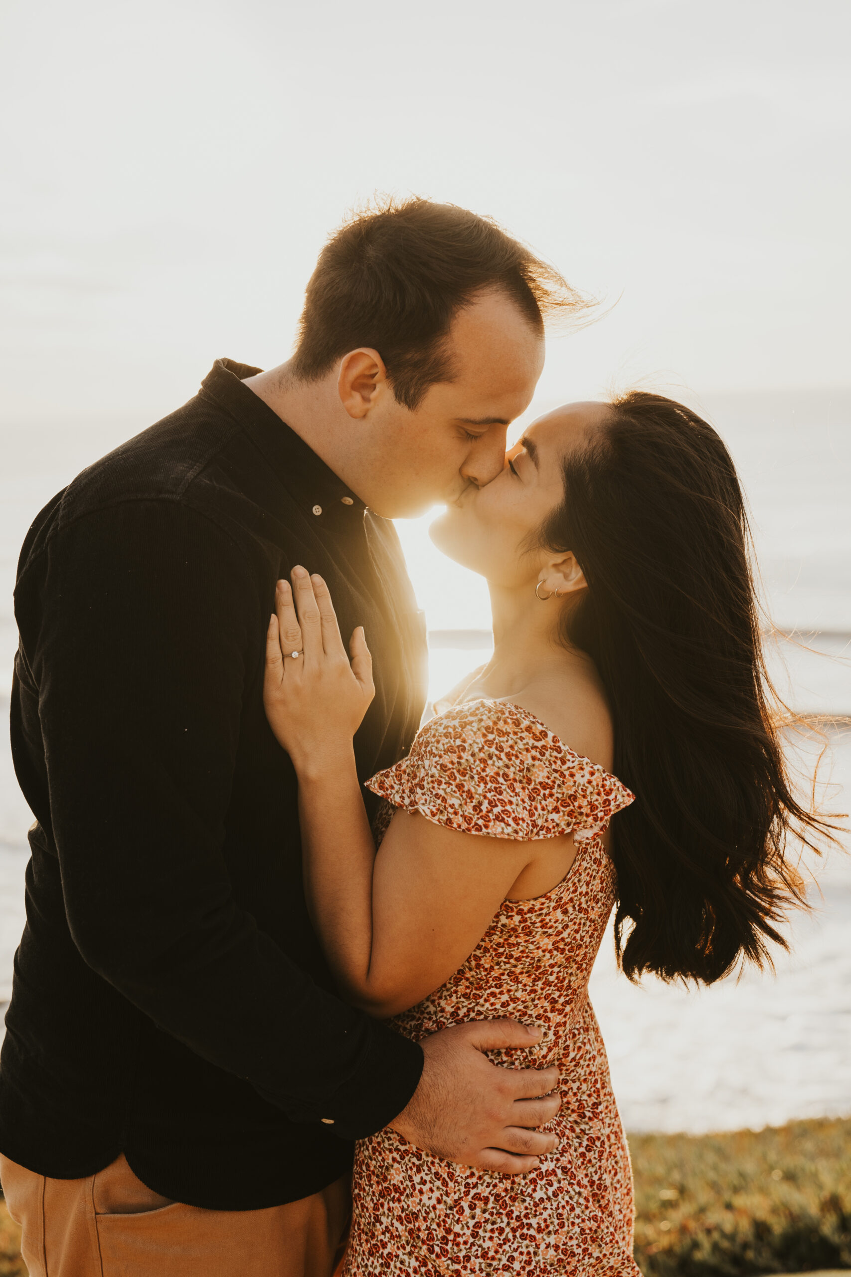 A couple stands wrapped up in each others arms, sun behind them, locked in a kiss with the Pacific Ocean sprawling out behind them.