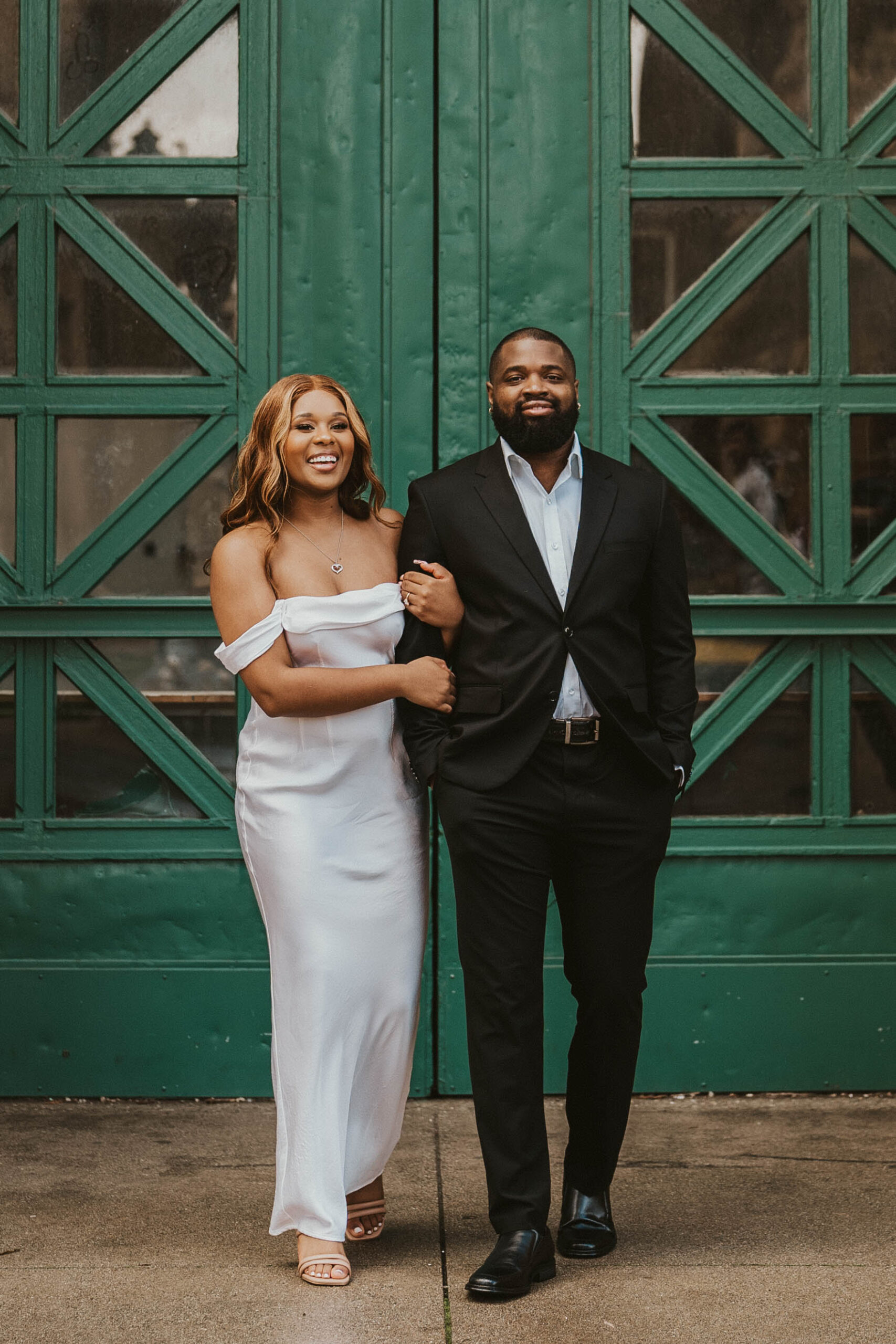 A couple walks happily towards the camera, arm in arm, laughing and smiling against a detailed deep green door. The woman is wearing a white satin gown and the man is wearing a stylish black suit.