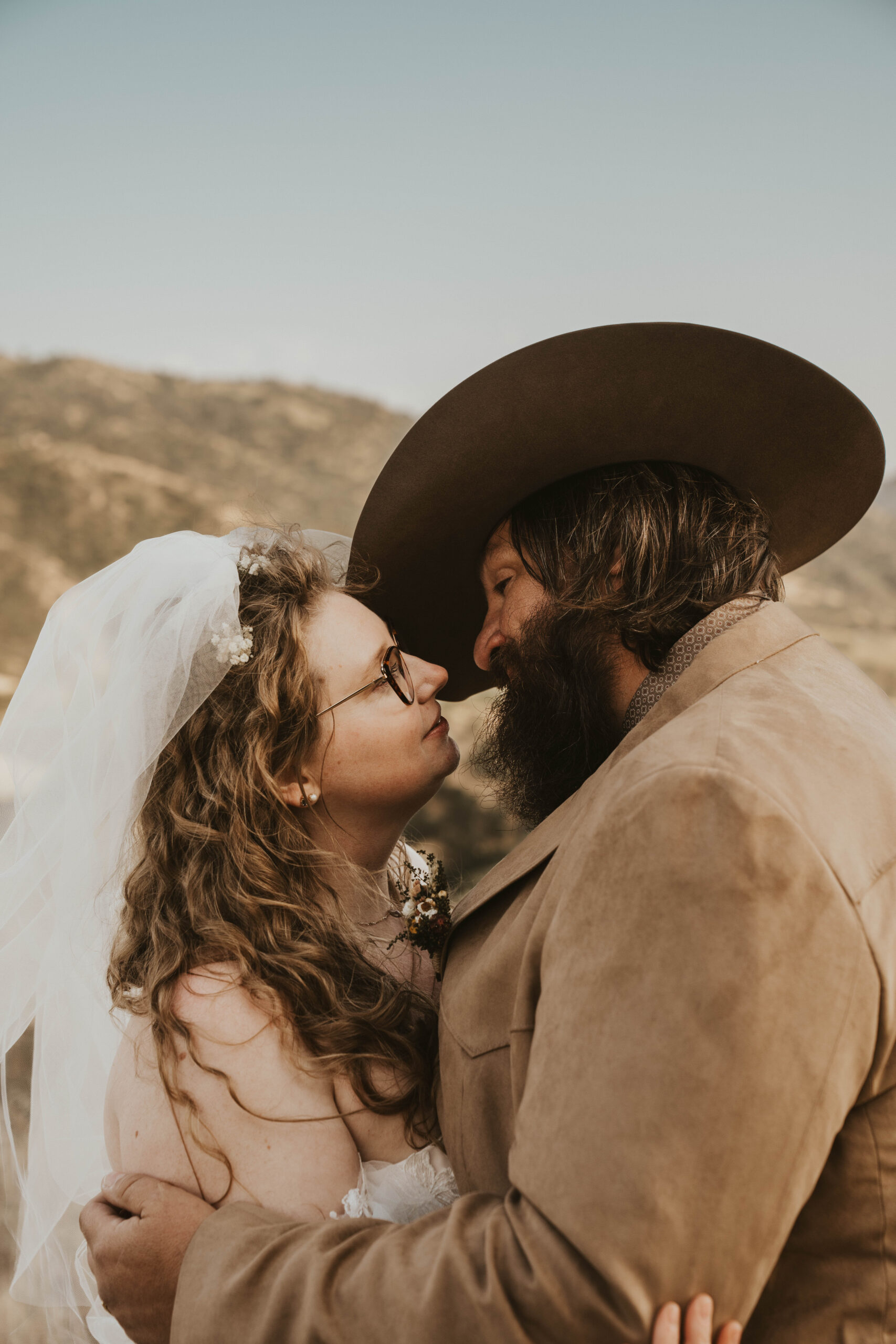 A couple stands close together atop a hill surrounded by rolling hills at Golden Hour.